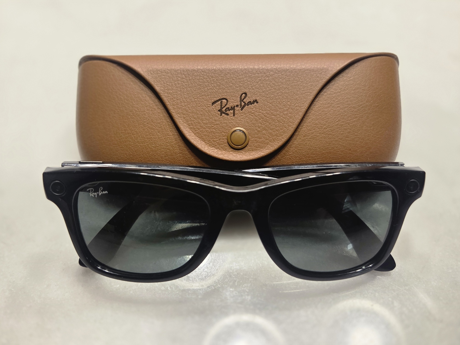 A pair of black Ray-Ban Meta smart glasses in front of their brown leather charging case