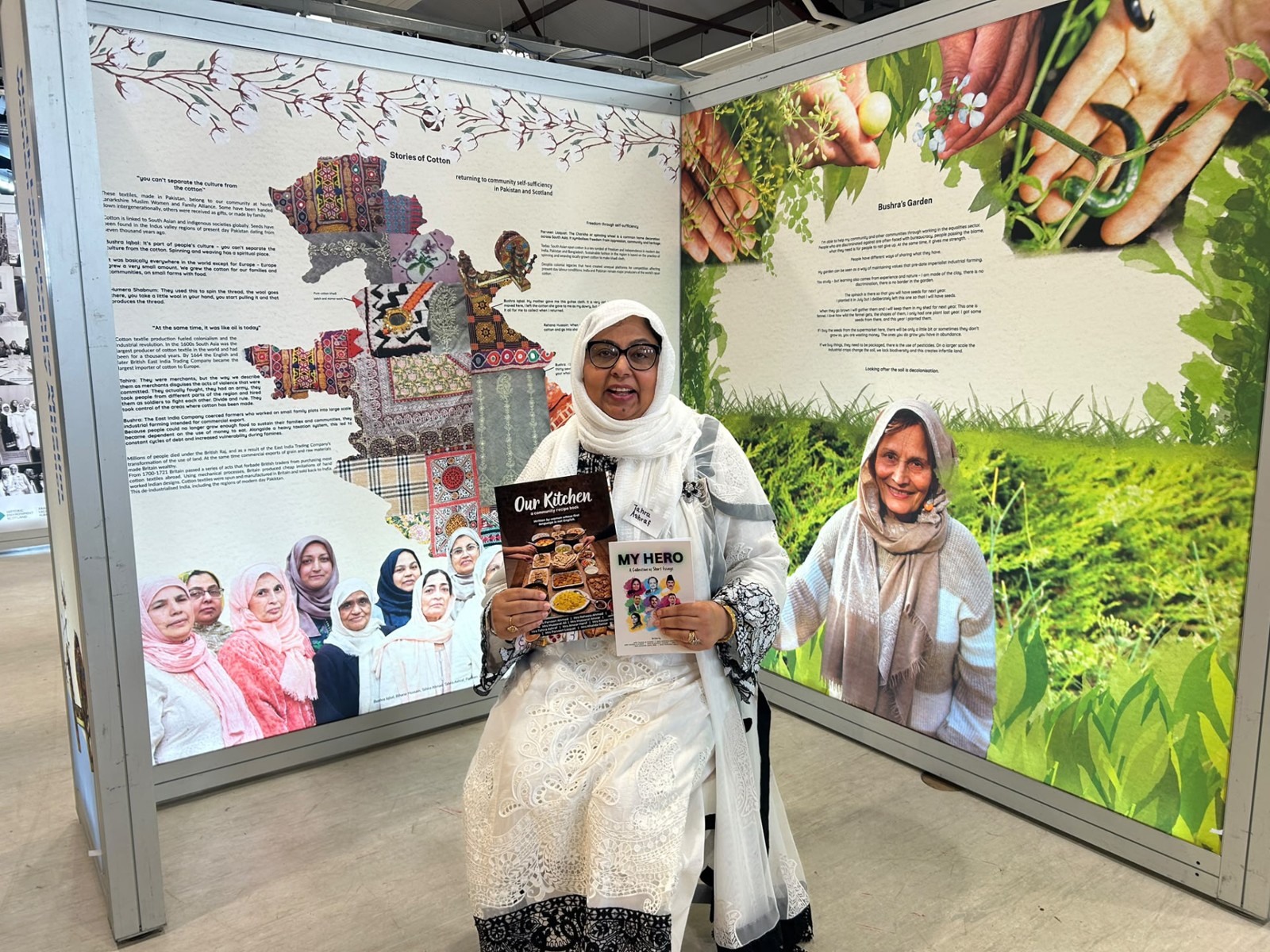 Exhibition participant holding Our Kitchen and My Hero books beside the Stories of Cotton and garden displays